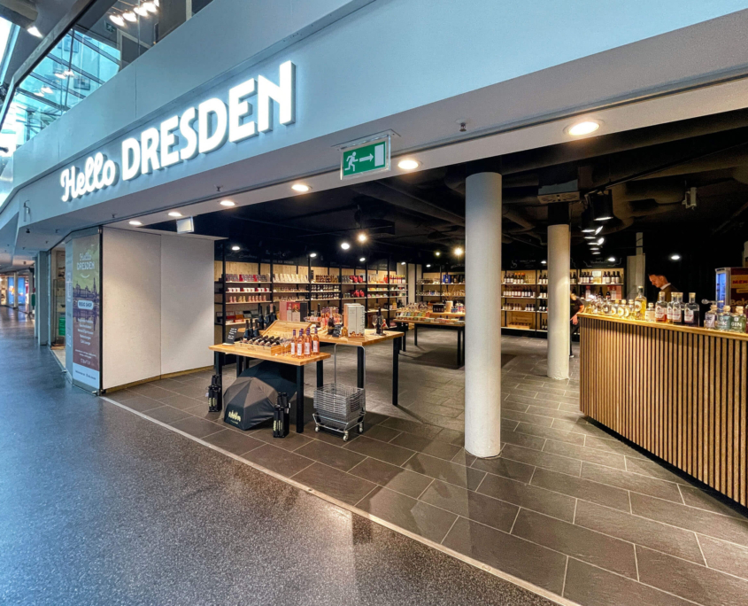 Hello-Dresden-Ladenansicht-01 Außenansicht Hello Dresden in der QF Passage mit Leuchtbuchstaben über Eingang, im Hintergrund Auslage mit Souvenirs und Ausschank zur Verkostung
